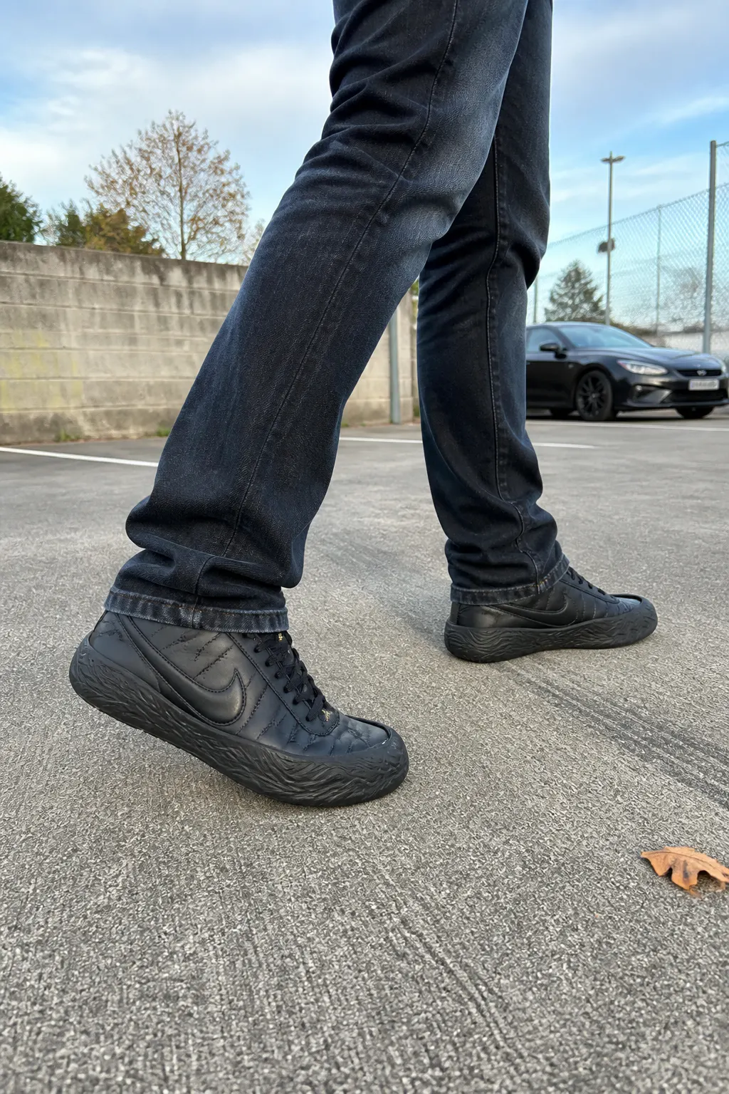 A footballer wearing studded boots with jeans on a pavement, matte-black CleatShield fitted, gripping the concrete