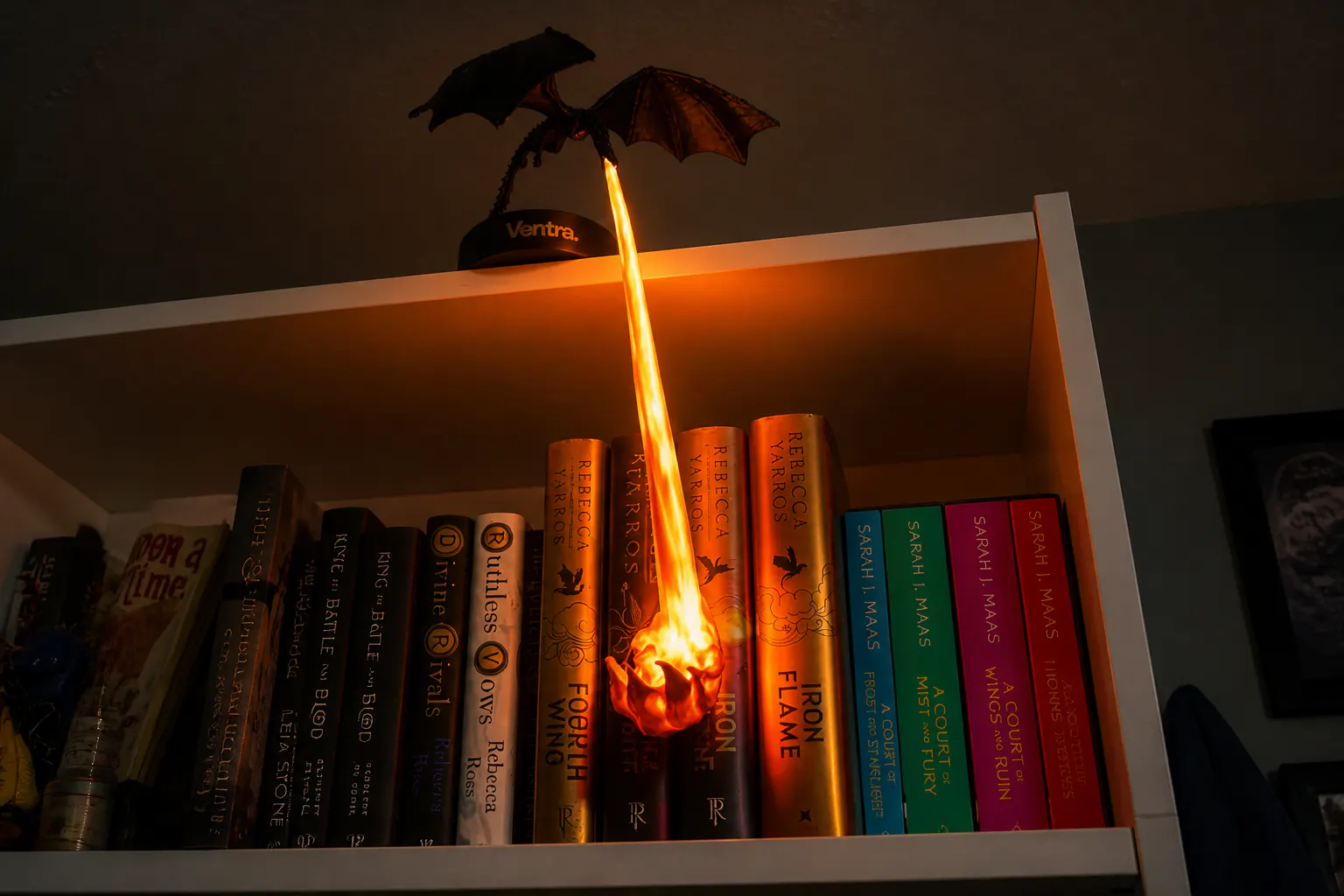 Lit dragon guardian breathing fire over bookshelf spines in a dim room
