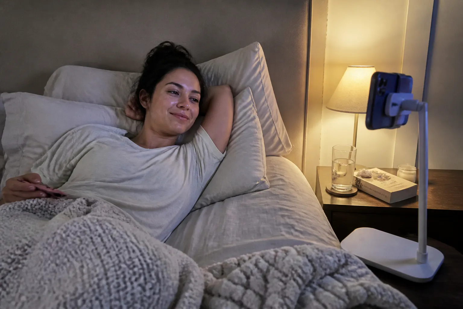 Dim warm bedroom, phone glowing in the ScrollMate stand at eye height, a person fully reclined with only the pink remote in a relaxed hand