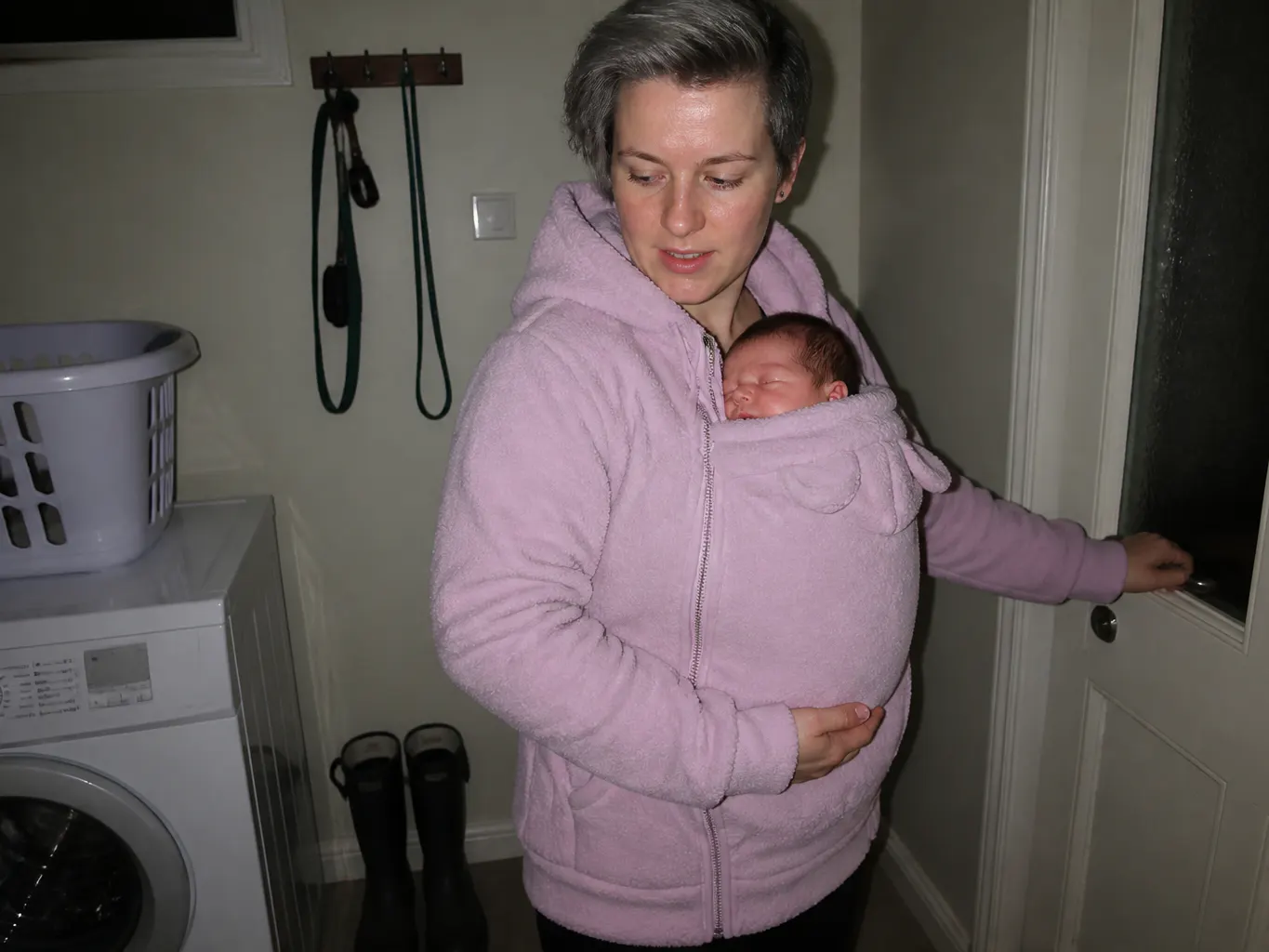 Parent in the fleece hoodie under a rain shell on a wet doorstep, baby in the pouch